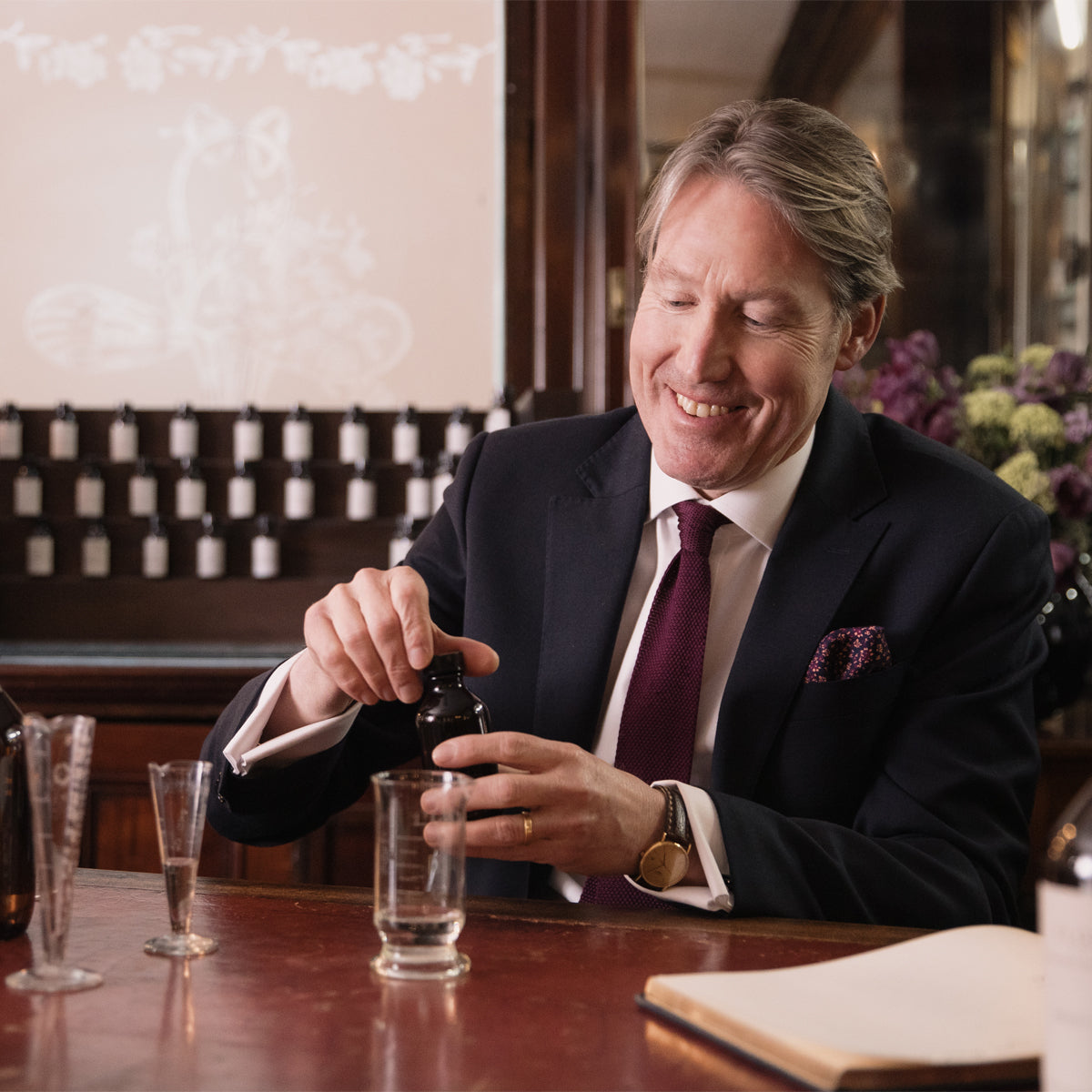 A smiling man in a suit mixes ingredients for a bespoke 100ml Fragrance Customisation by Floris London at a table with glassware and a notebook.