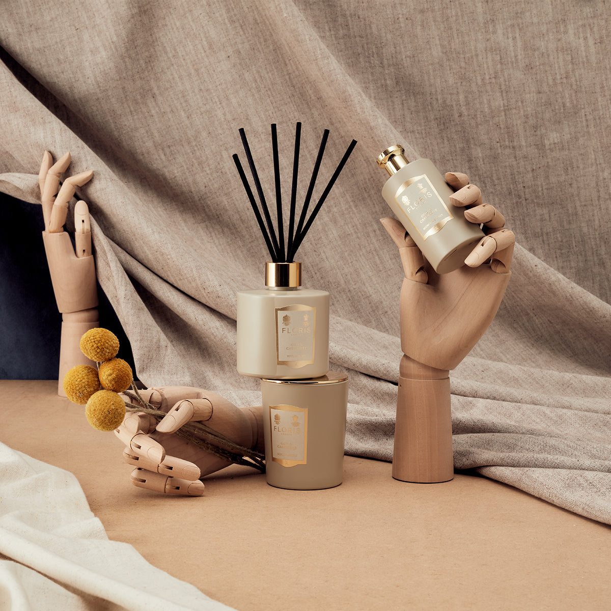 Three wooden mannequin hands display Floris London's Oud & Cashmere Reed Diffuser, scented candles, and yellow billy ball flowers on a neutral backdrop.