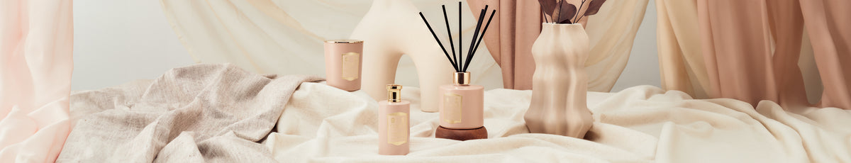 Elegant perfume bottles and scented reed diffuser on a soft, draped fabric backdrop with warm, neutral tones.