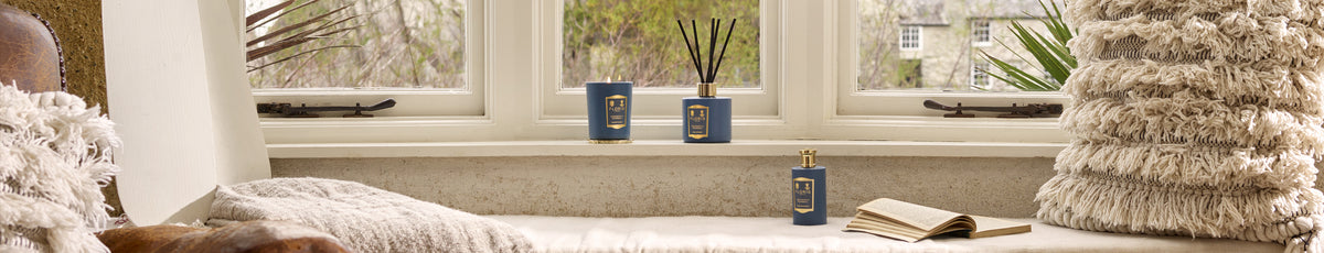 Candle, reed diffuser, and book on a cozy window seat with fluffy cushions and a view of greenery outside.