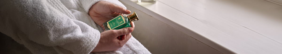 Person in a white robe holding a bottle of perfume with a green label, near a windowsill.
