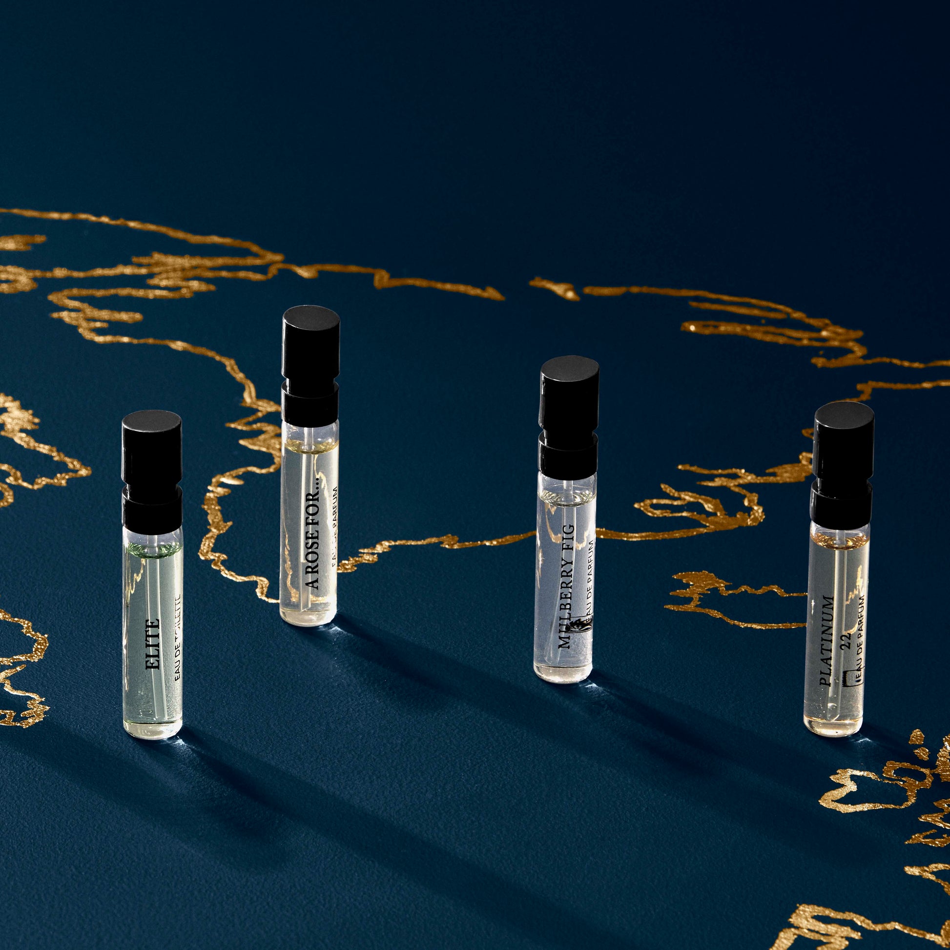 Four Floris London Sample Set vials stand on a blue, gold-patterned surface—includes $25 off any 50ml or 100ml fragrance purchase.