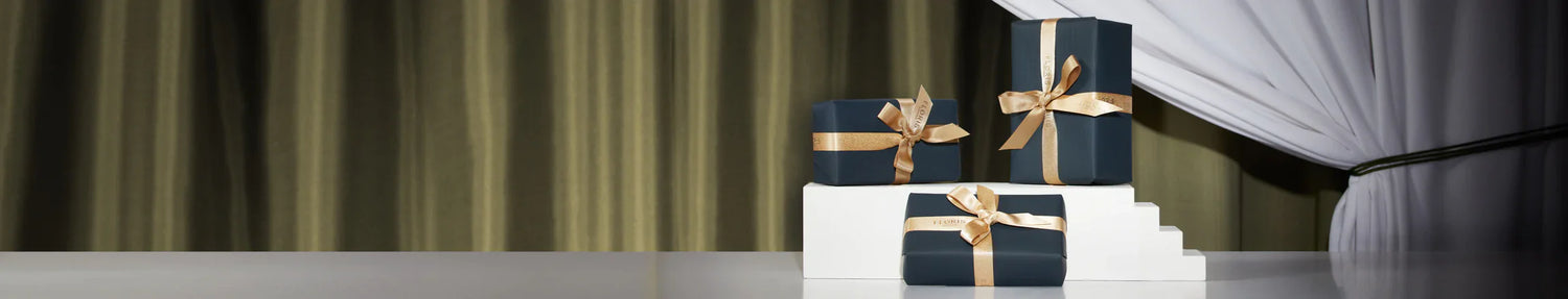 Three wrapped presents with gold bows stacked together on a reflective surface in front of green curtains and white drapes.
