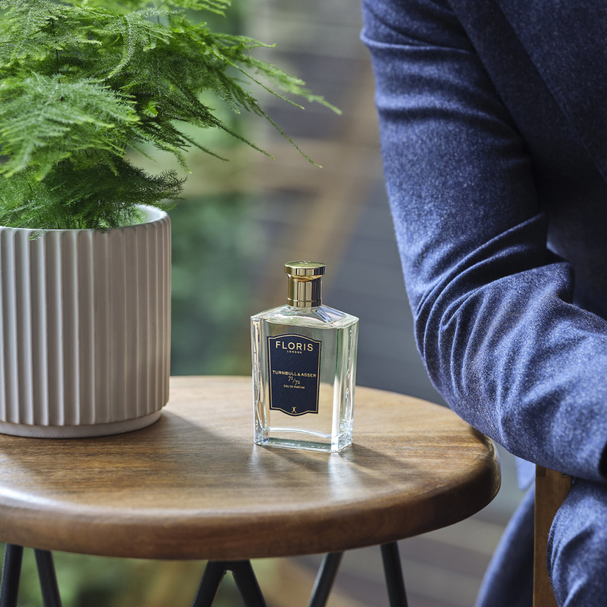 71/72 - Eau de Parfum by Floris London rests on a wooden table next to someone in a blue jacket and a potted plant.