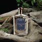 A clear glass bottle of Floris London 71/72 - Eau de Parfum rests on driftwood with green plants and pebbles—a refined woody fragrance.