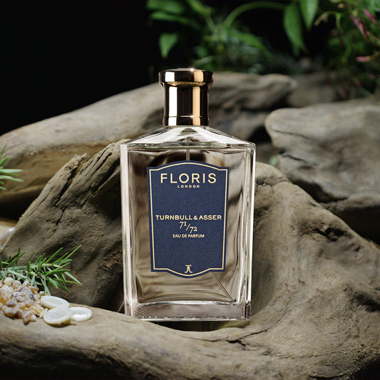 A clear glass bottle of Floris London 71/72 - Eau de Parfum rests on driftwood with green plants and pebbles—a refined woody fragrance.