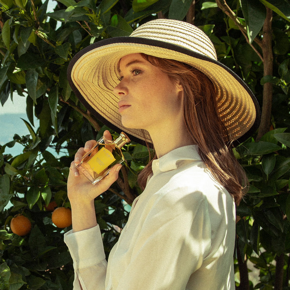 A woman in a wide-brimmed hat holds Bergamotto di Positano—an Eau de Parfum by Floris London—near orange trees, surrounded by the scent of orange blossom.