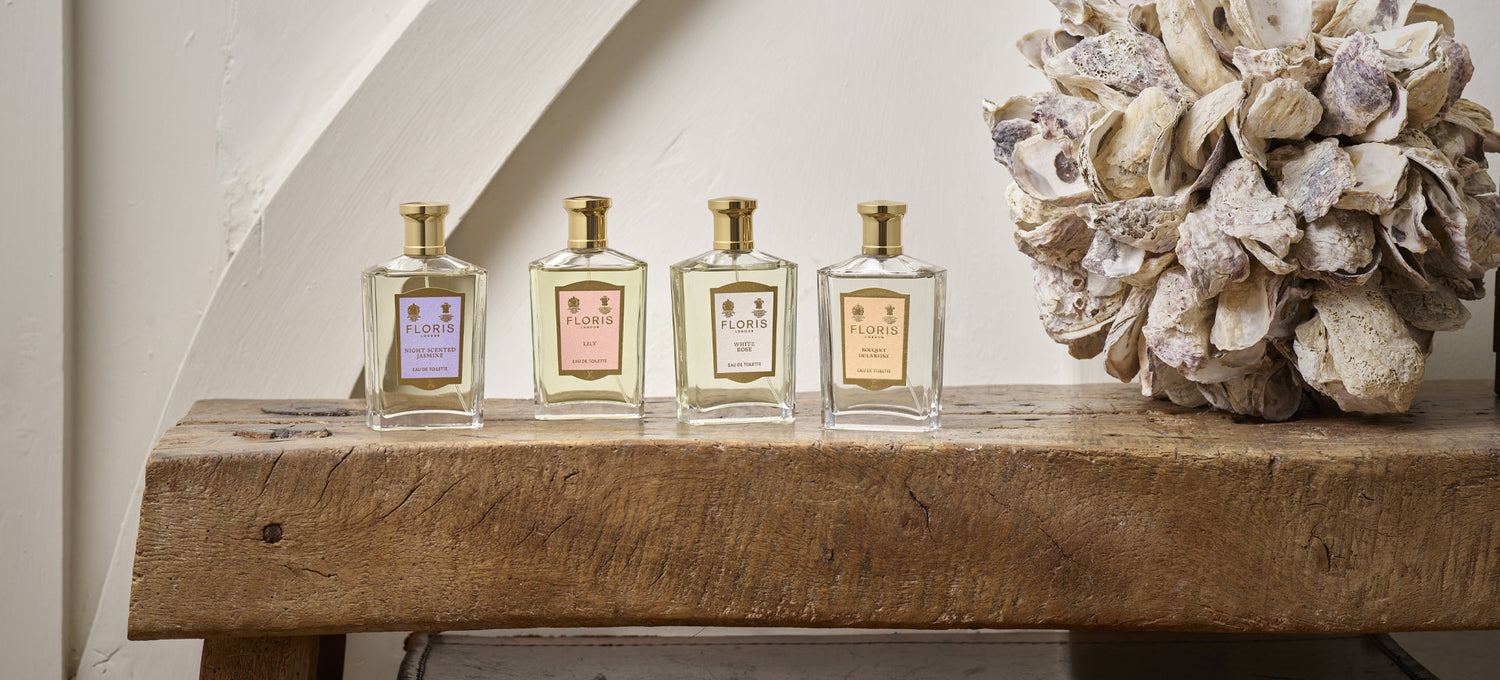 Four perfume bottles with gold caps and various labels on a rustic wooden bench beside a decorative shell arrangement.