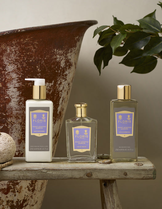 The Floris London Night Scented Jasmine Bath & Body Collection placede upon a woden milking stool nex to a vintage bathtub. 