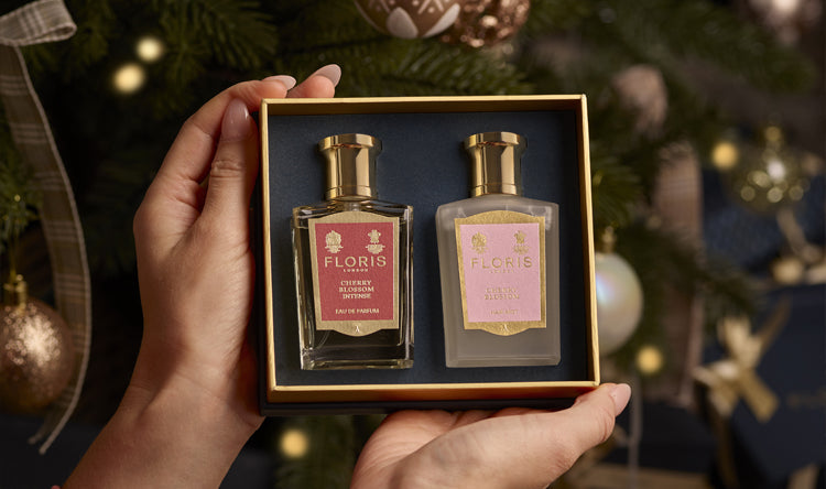 Hands holding a boxed Cherry Blossom Duet from Floris London, featuring two exquisite perfumed products, in front of a Christmas tree adorned with ornaments and lights. This luxurious gift set is perfect for the holiday season.