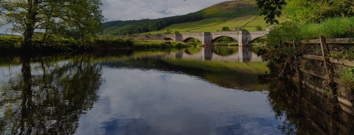 Stone bridge over a calm river with lush green hills and trees reflecting in the water under a partly cloudy sky.