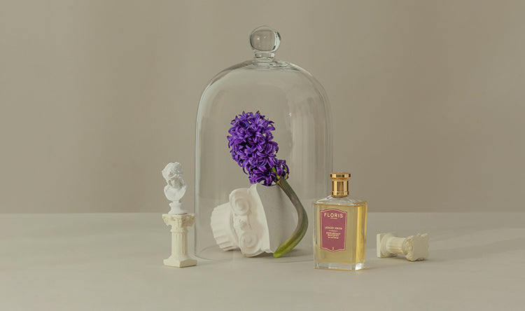 Purple flowers under a glass dome, surrounded by white bust sculptures, with a bottle of perfume labeled "Floris" beside them.