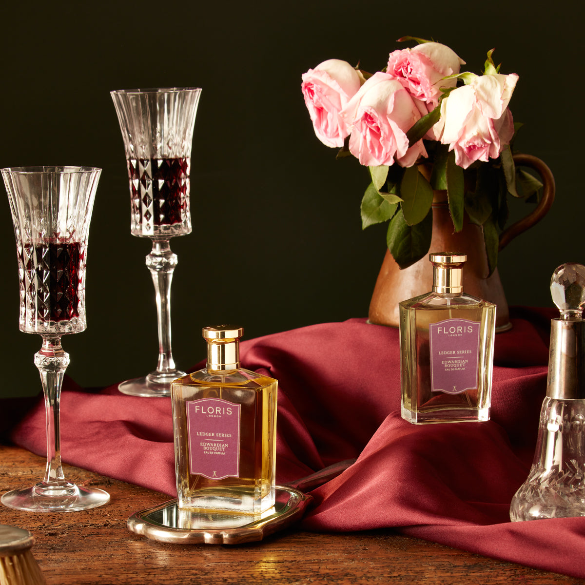 Elegant scene featuring Floris London Edwardian Bouquet with wine glasses, pink roses in a vase, and burgundy fabric on a wooden table.