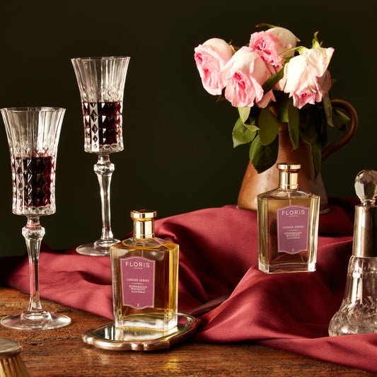 Elegant scene featuring Floris London Edwardian Bouquet with wine glasses, pink roses in a vase, and burgundy fabric on a wooden table.