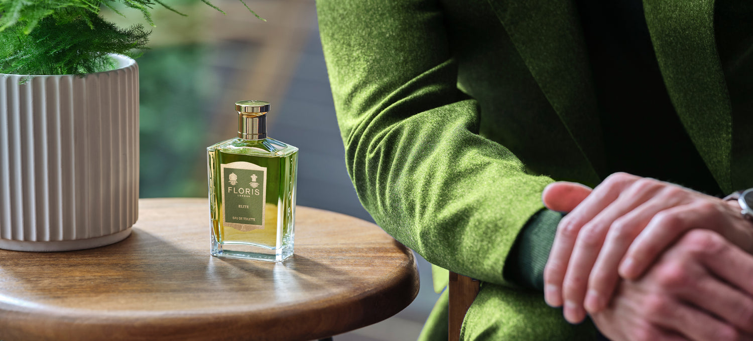 A bottle of Elite on a wooden table with a potted fern, and a man in a green tweed suit leaning on the table.