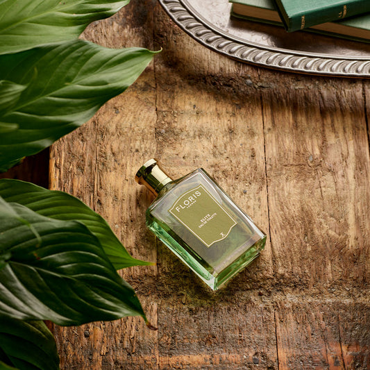A bottle of Floris London Elite - Eau de Toilette sits on rustic wood with citrus leaves and books, evoking a fresh, woody ambiance.