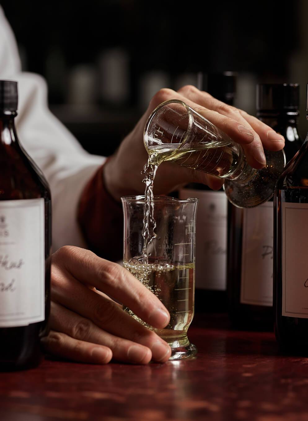 Person pouring liquid from a small flask into a beaker surrounded by dark bottles with labels, on a wooden table.