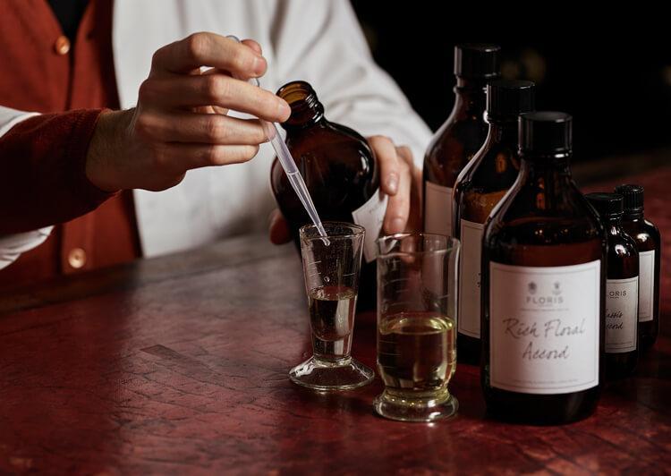 Person in white coat using a dropper to add liquid to a glass, with various amber bottles on the table.