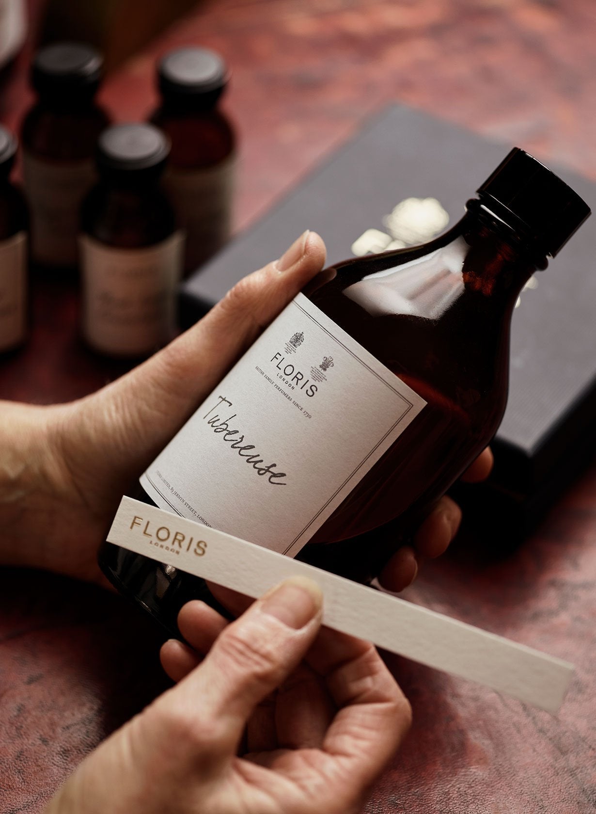 Person holding a Floris fragrance bottle and a scent strip labeled "FLORIS" with more small bottles in the background.