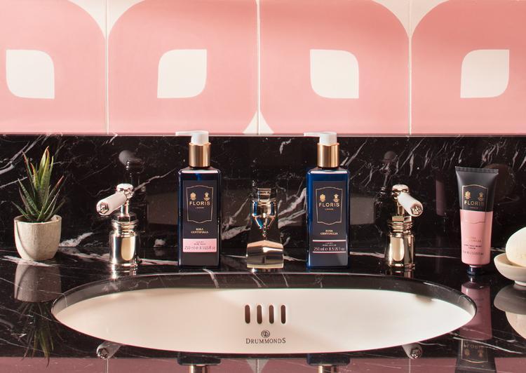 Modern bathroom sink with soaps, lotions, and plant on a black marble counter with pink and white tiled backsplash.