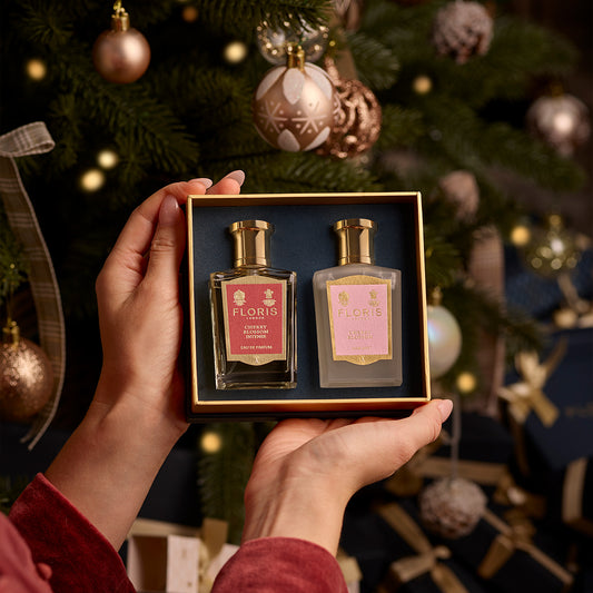 Hands holding the exquisite Cherry Blossom Duo from Floris London, its delicate fragrance perfectly complementing the festive intensity of a beautifully decorated Christmas tree.