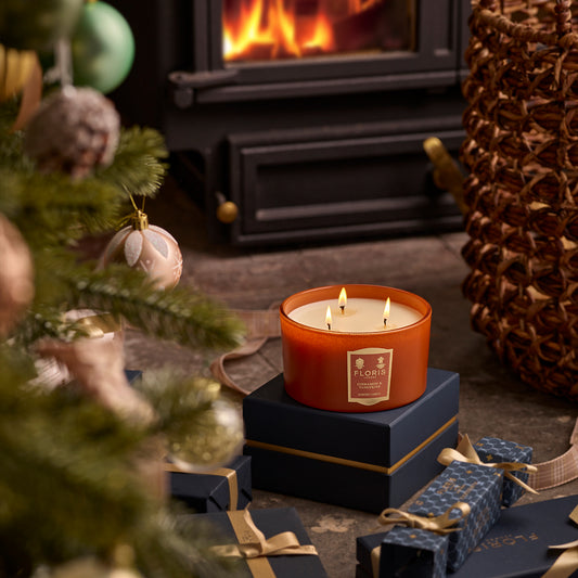 Lit a Floris London Cinnamon & Tangerine Deluxe Three Wick Candle on the gift boxes near the decorated Christmas tree and fireplace, filling the air with its limited-edition aroma and creating a cozy holiday atmosphere.