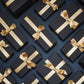 Numerous black gift boxes with gold ribbons arranged in neat rows on a dark background.