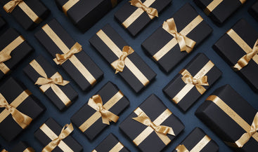 Numerous black gift boxes with gold ribbons arranged in neat rows on a dark background.