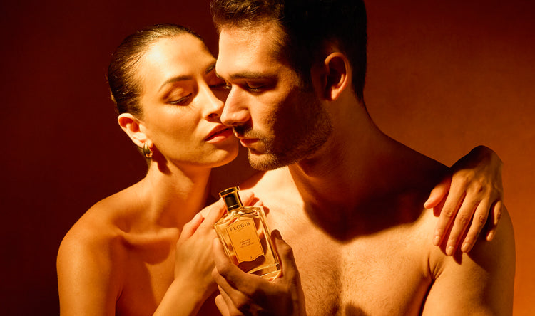 A woman embraces a shirtless man and holds a perfume bottle close to his face with a warm lit background.