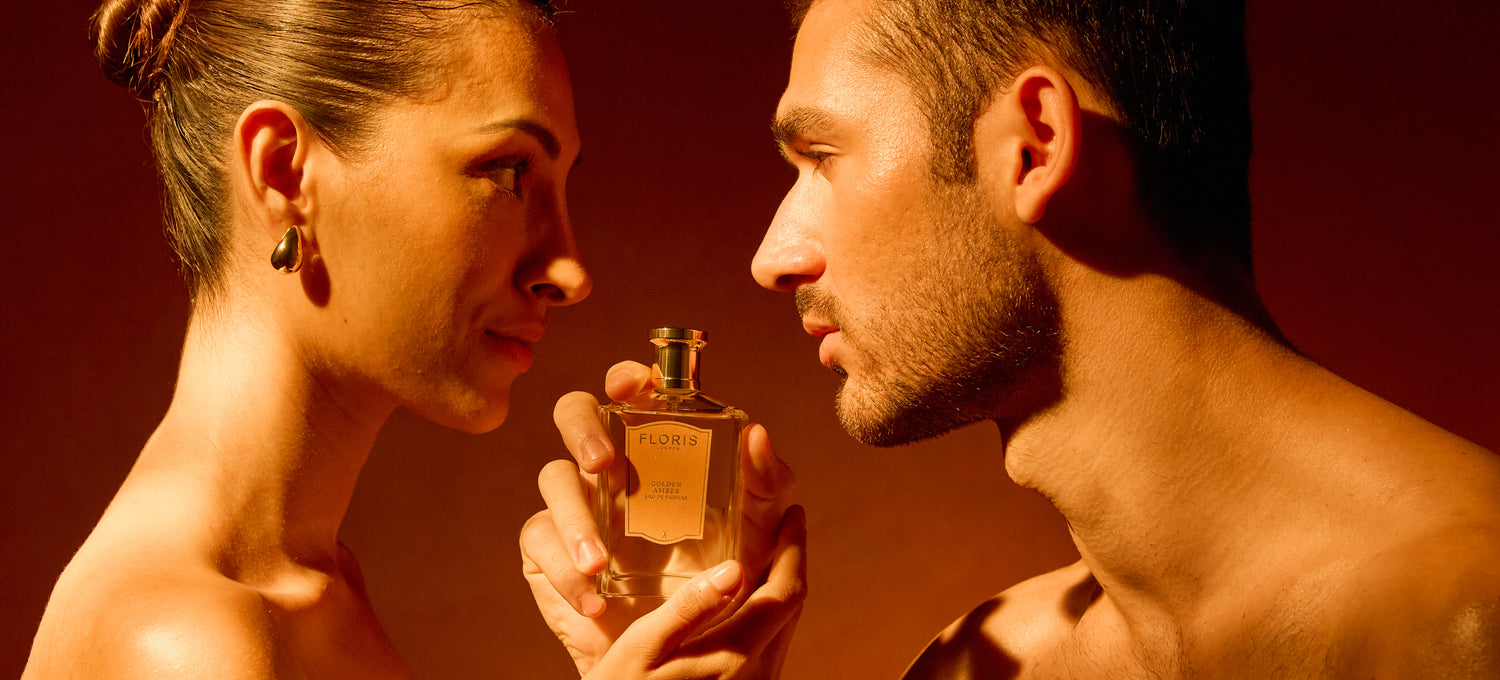 Two people face each other closely, holding a Floris perfume bottle between them against an amber background.