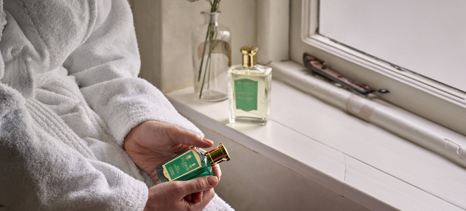 A woman in a bath robe, holding a bottle of Floris London Roe Geranium Bath Essence next to a window ledge with Rose Geranium Eau de Parfum. 