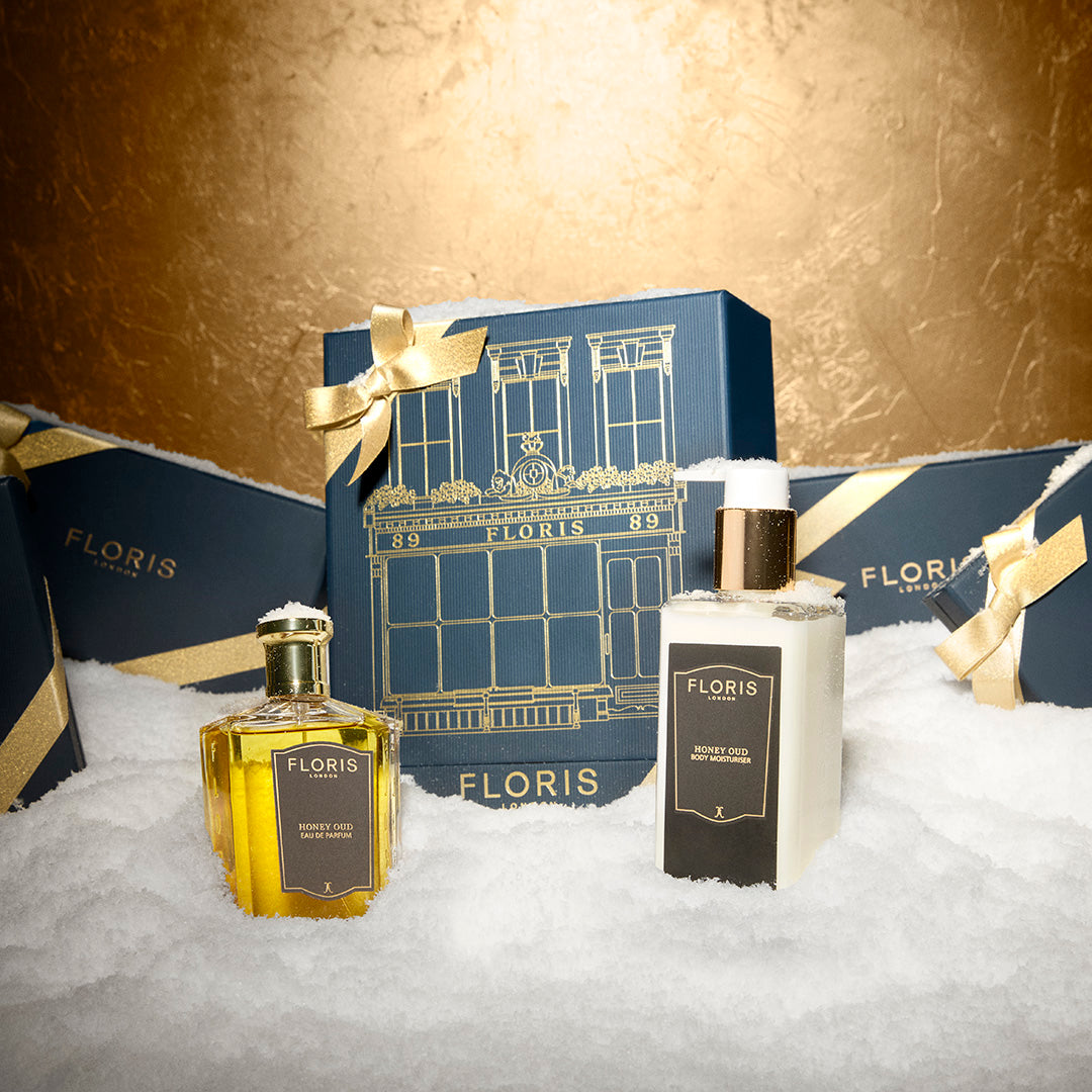 Two Floris London UK Sweet Lux | Honey Oud Duo bottles with a blue gift box and gold ribbon are elegantly displayed on artificial snow.