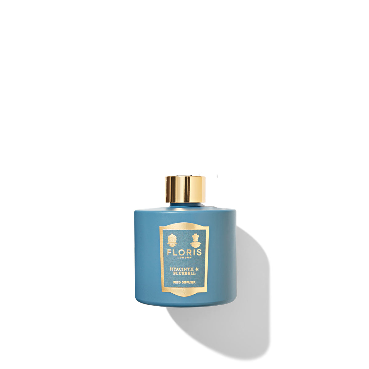 A Hyacinth & Bluebell - Reed Diffuser by Floris London, featuring a blue bottle with a gold cap and label, casts a soft shadow on a white background while filling the space with a delicate floral fragrance reminiscent of spring flowers.