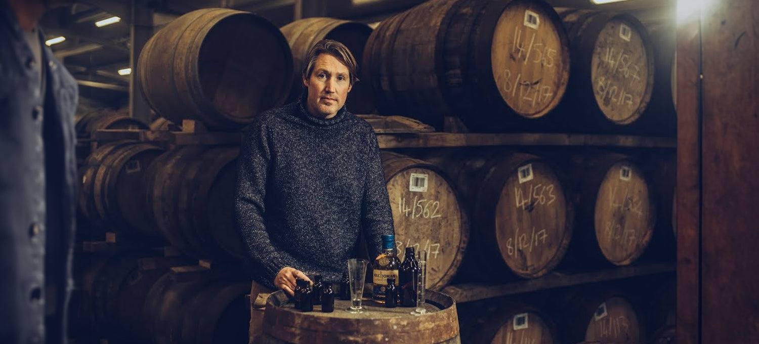 A person stands by a barrel with whiskey bottles and glasses in an aging warehouse filled with stacked barrels.