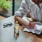 A man writes at a wooden table with JF - Eau de Toilette by Floris London, boasting citrus and woody notes, next to a pair of glasses.