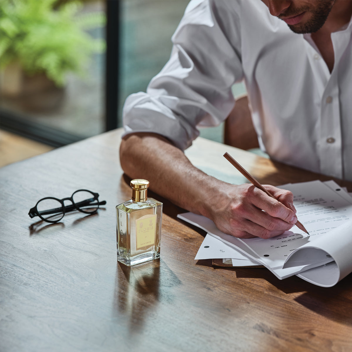 A man writes at a wooden table with JF - Eau de Toilette by Floris London, boasting citrus and woody notes, next to a pair of glasses.