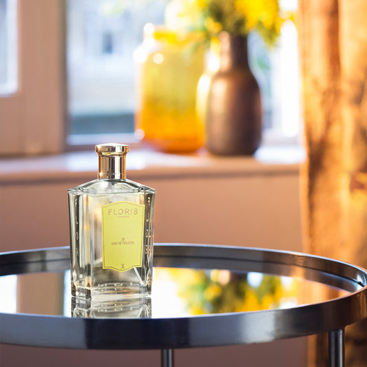 A Floris London JF - Eau de Toilette bottle rests on a glass table, vases and flowers blurred behind; a citrus woody fragrance for men.