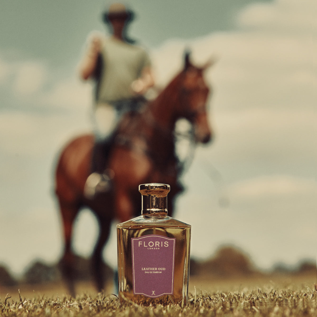 A Floris London Leather Oud Eau de Parfum bottle with a blurry rider in the background, outdoors. Captivatingly blends vetiver and oud oil.