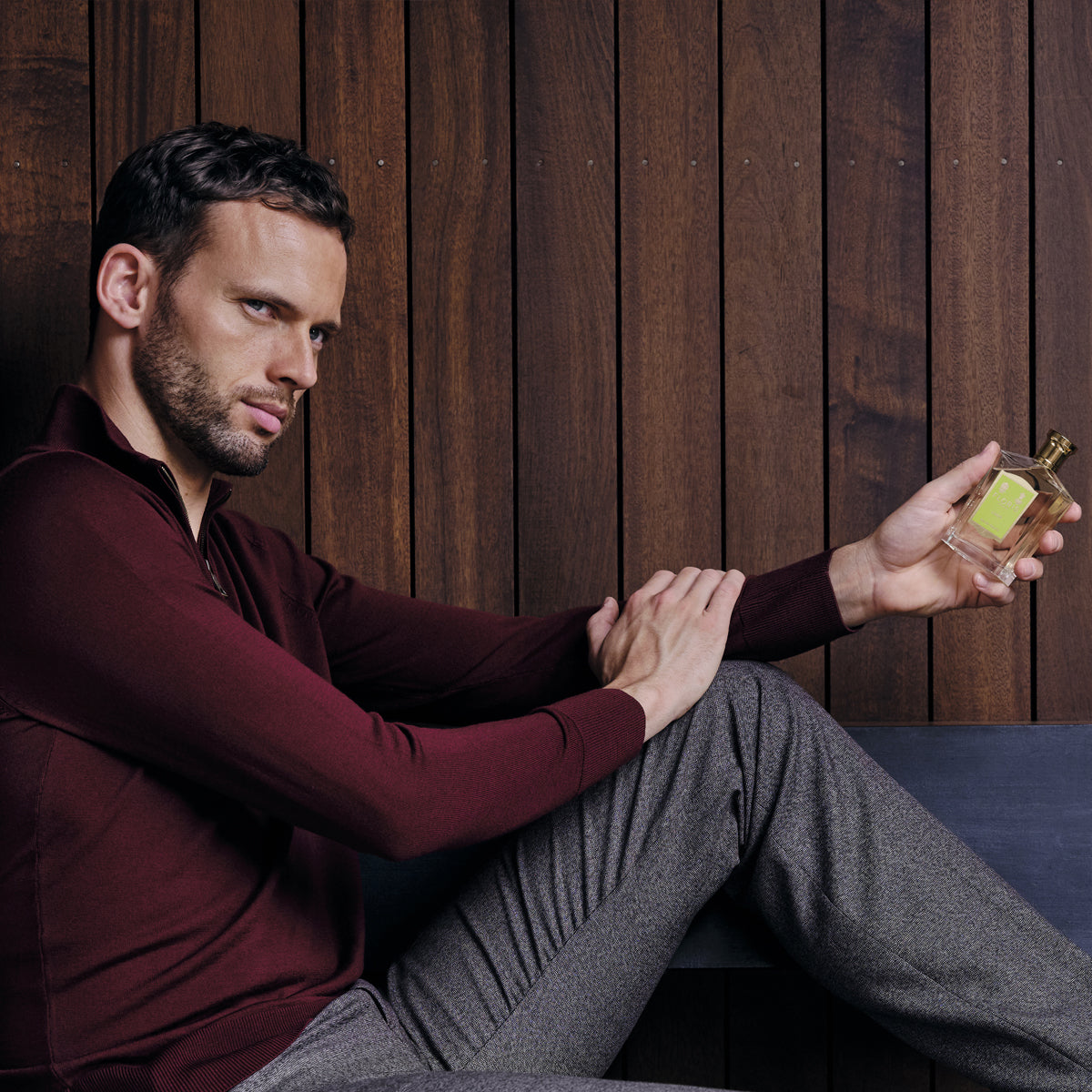 A man in a red sweater and gray pants sits against a wooden wall, holding Floris London's Limes - Eau de Toilette with a contemplative expression.