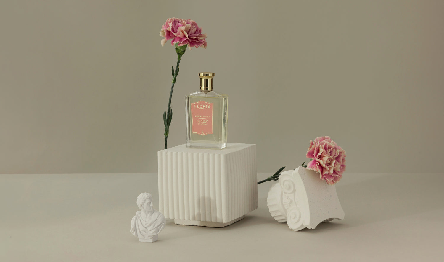 A perfume bottle with a pink label on a white pedestal, surrounded by pink carnations and white decorative sculptures.