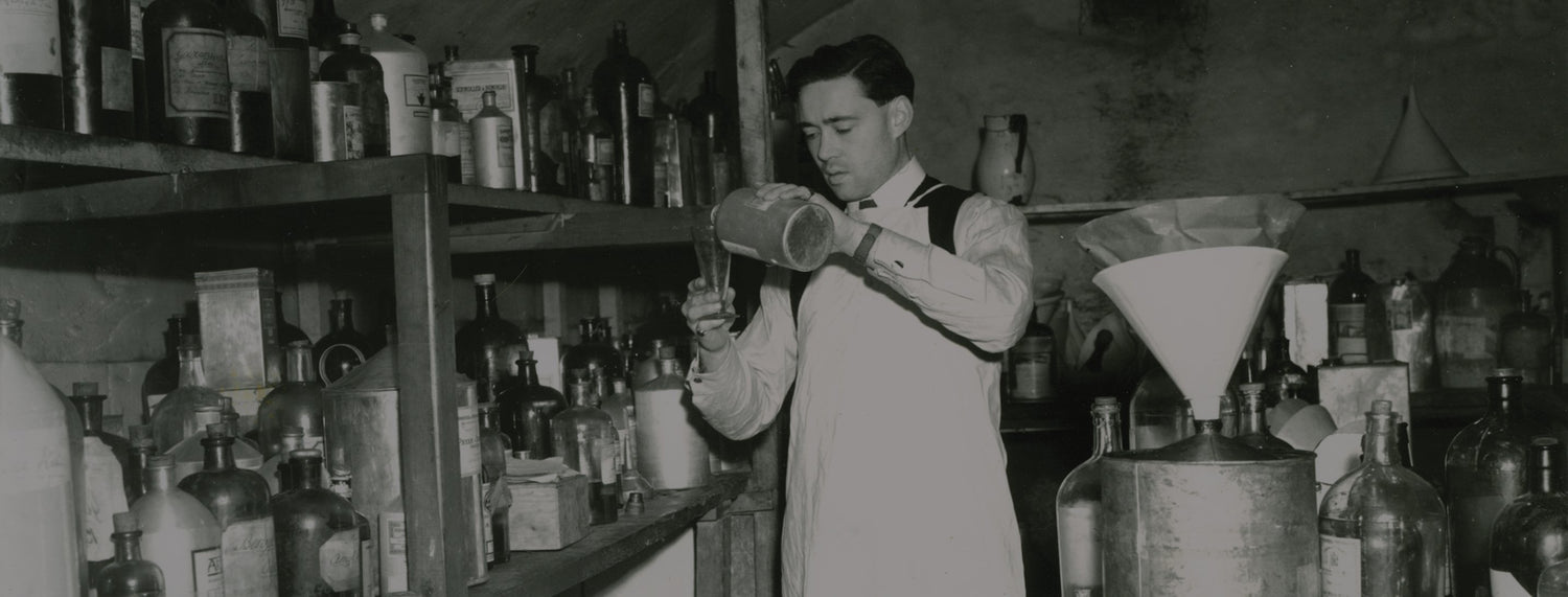 A man in a lab coat pouring liquid into a beaker in a cluttered laboratory filled with bottles and equipment.