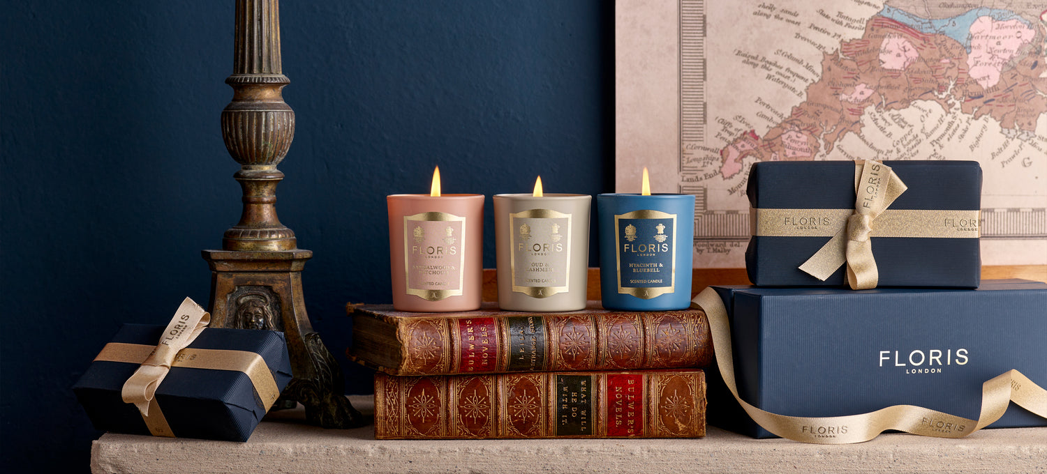 Three beautifully scented candles from the Miniature Candle Collection by Floris London sit on antique books near a decorative map, beside gift-wrapped boxes with ribbons on a mantel.