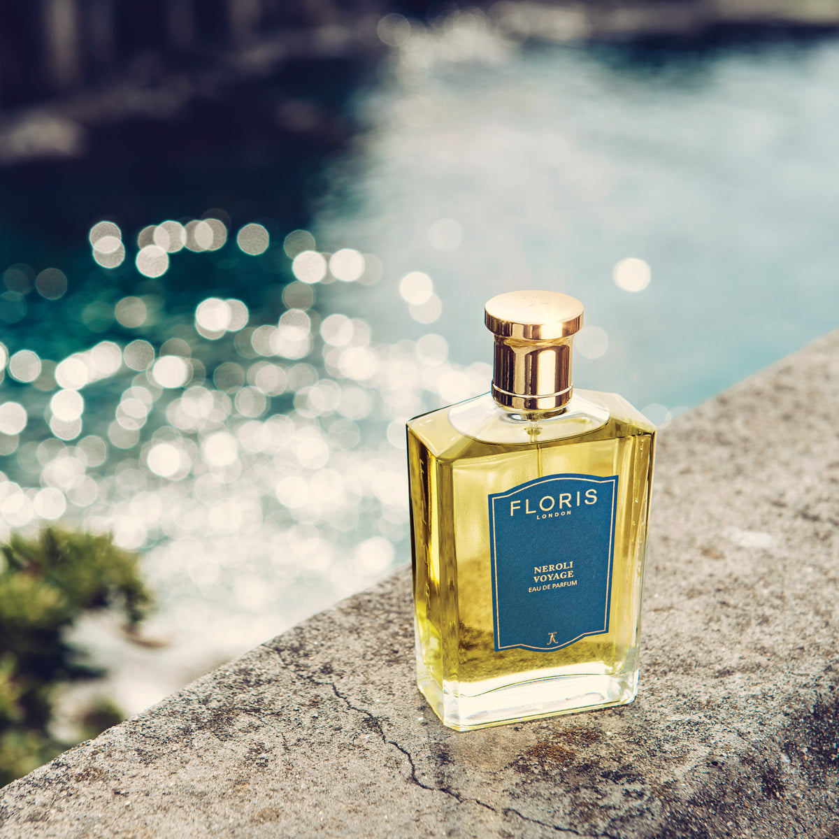 Neroli Voyage - Eau de Parfum by Floris London sits on a ledge with a blue label, overlooking sunlit waters and capturing a fresh citrus allure.