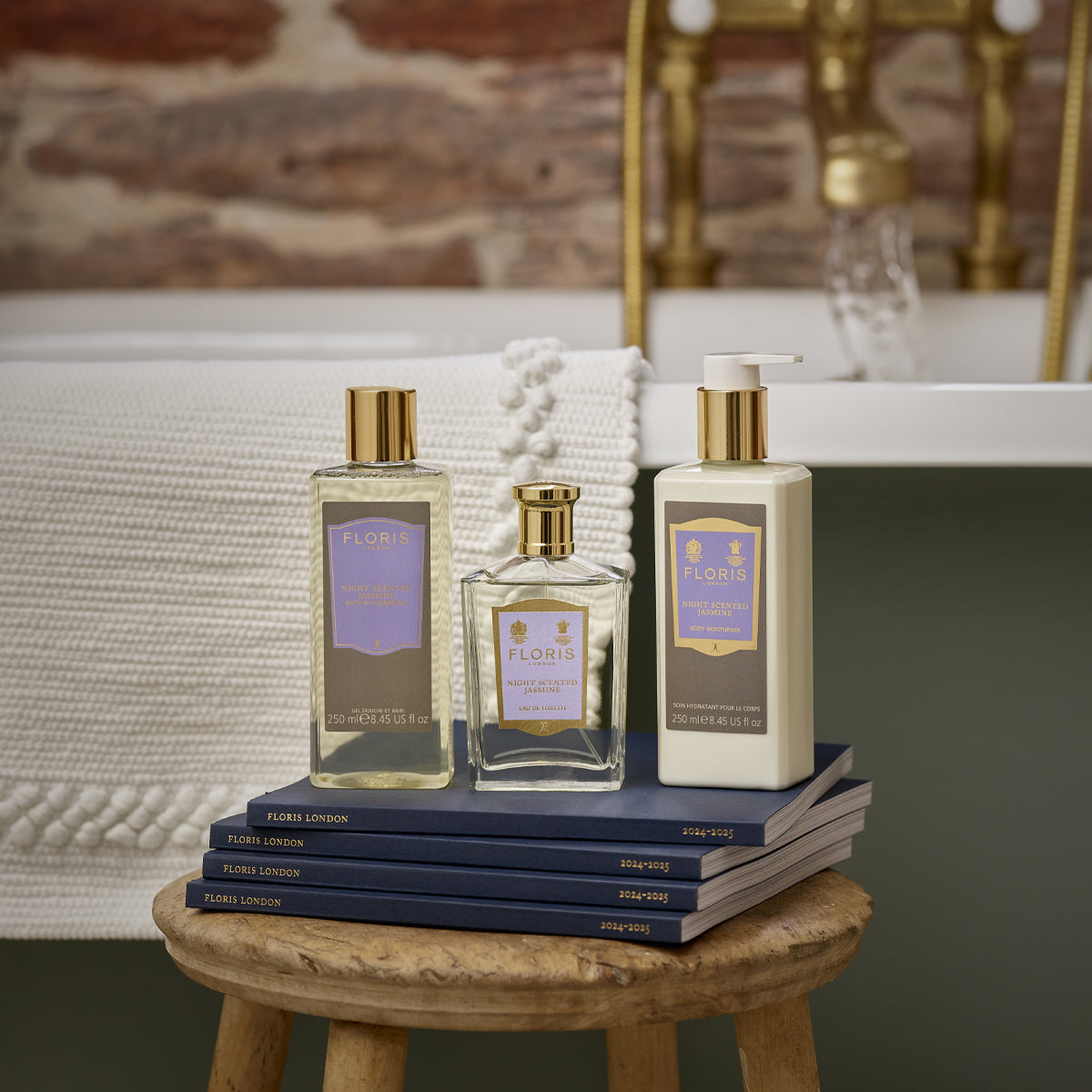 Three Floris London products, their Night Scented Jasmine aroma wafting through the air, sit on stacked books before a tub draped with a towel.