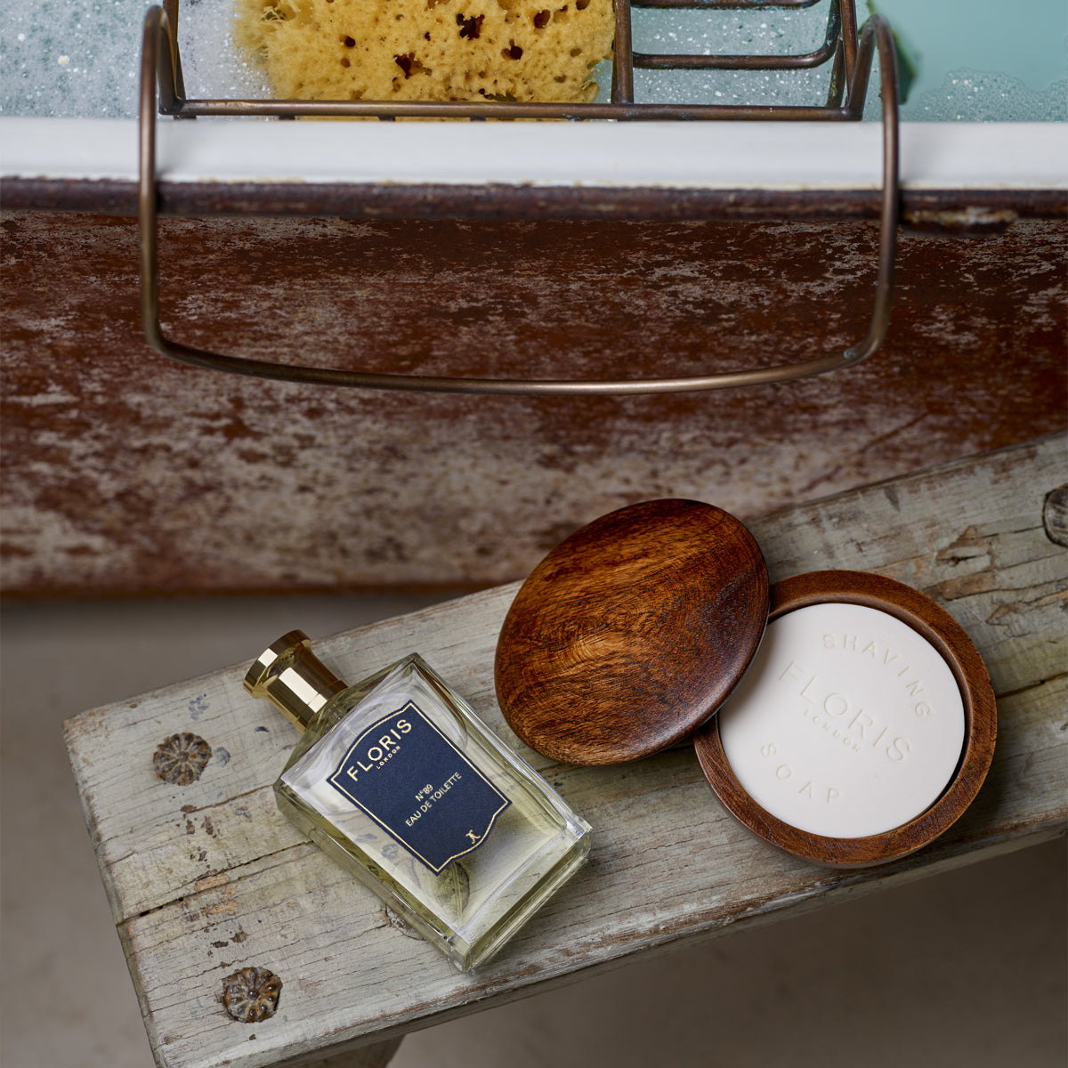 Wooden bench with Floris London's No. 89 Eau de Toilette, vegan shaving soap, and a sponge near a bubble-filled bathtub.