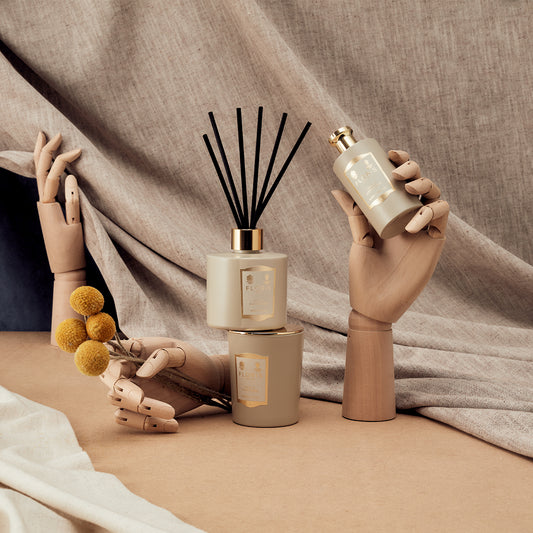 Three wooden mannequin hands display Floris London's Oud & Cashmere Reed Diffuser, scented candles, and yellow billy ball flowers on a neutral backdrop.