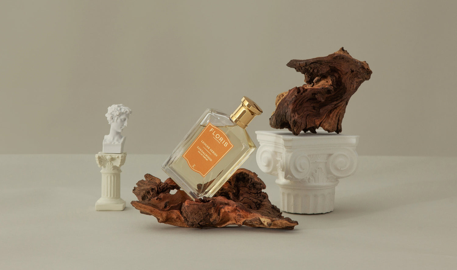 Perfume bottle on driftwood with mini classical statue, ornate column, and wood sculpture in a minimalist setting.