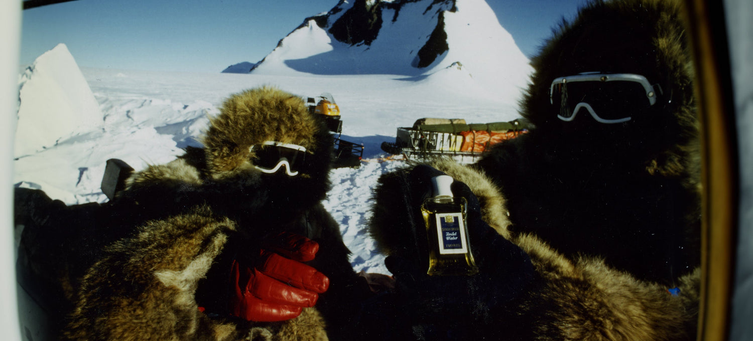 Two people in furry coats and goggles hold a bottle with mountains and a snow-covered landscape in the background.