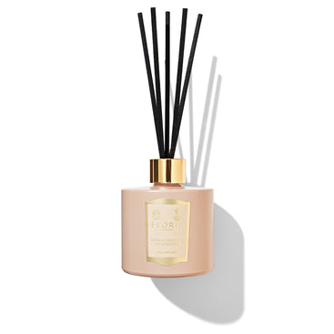 A pink Sandalwood & Patchouli reed diffuser by Floris London, featuring a gold label and black sticks and casting a shadow against a white background, fills the room with hints of sandalwood.