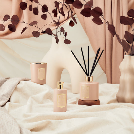 Floris London’s Sandalwood & Patchouli Reed Diffuser, pink bottles, and a candle rest on cream fabric with dried leaves and a beige backdrop.